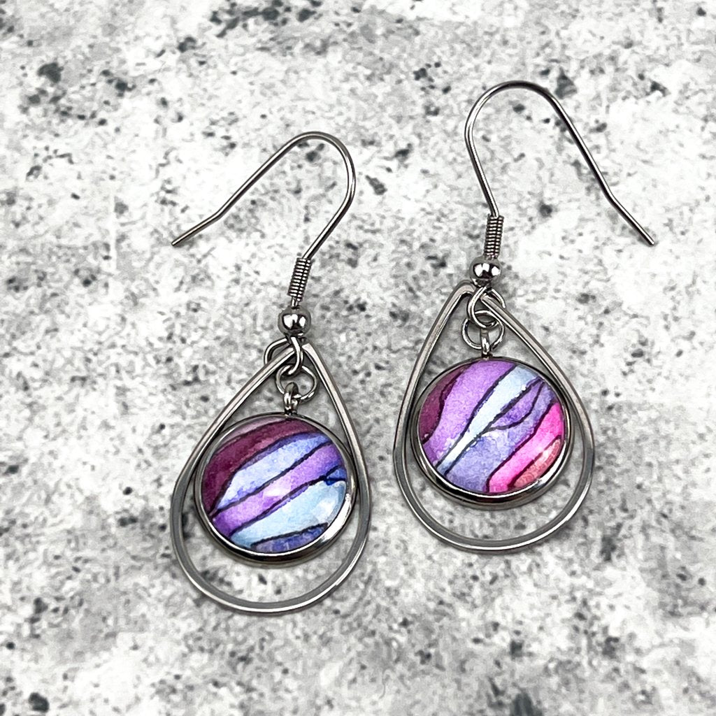Purple Rain dangle earrings, which feature a round glass cabochon overtop watercolor art in wavy ripples of purple paint. Art is surrounded by a teardrop shaped stainless frame. 