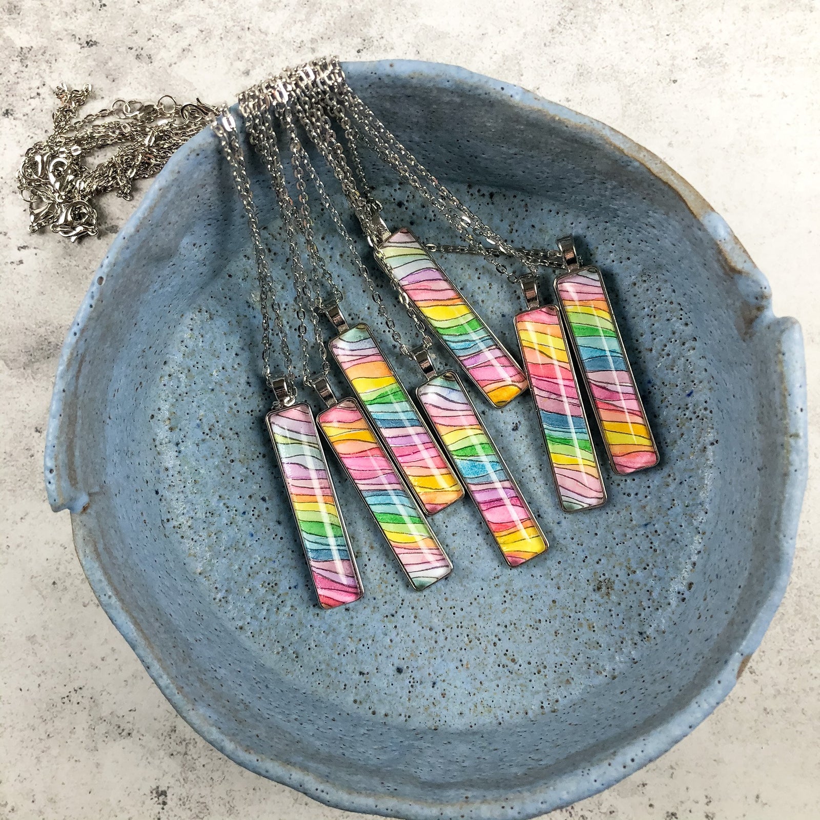 Narrow rectangular rainbow waves pendants displayed in a handmade blue ceramic bowl. 