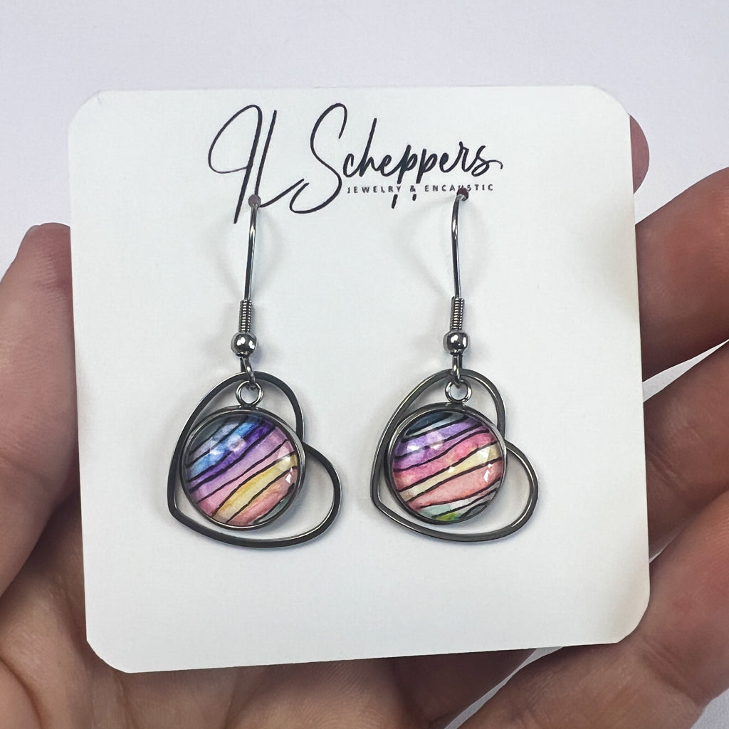 Drop earrings featuring a heart-shaped frame surrounding round rainbow-colored watercolor art behind glass. 