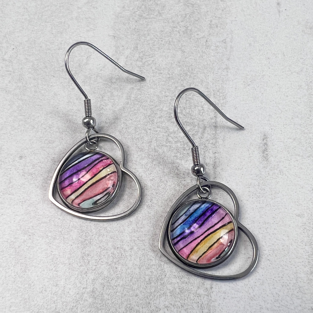 Drop earrings featuring a heart-shaped frame surrounding round rainbow-colored watercolor art behind glass. 