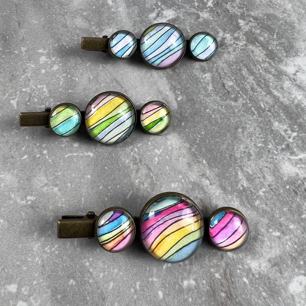 Three rainbow wave brass hair clips on a grey background. 