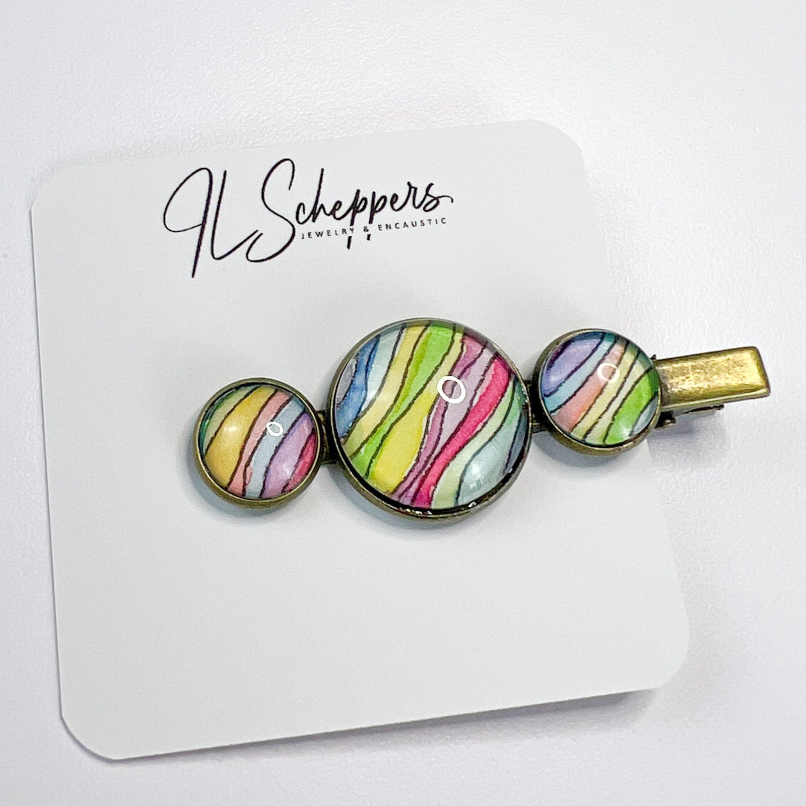 Three rainbow wave brass hair clips on a grey background. 