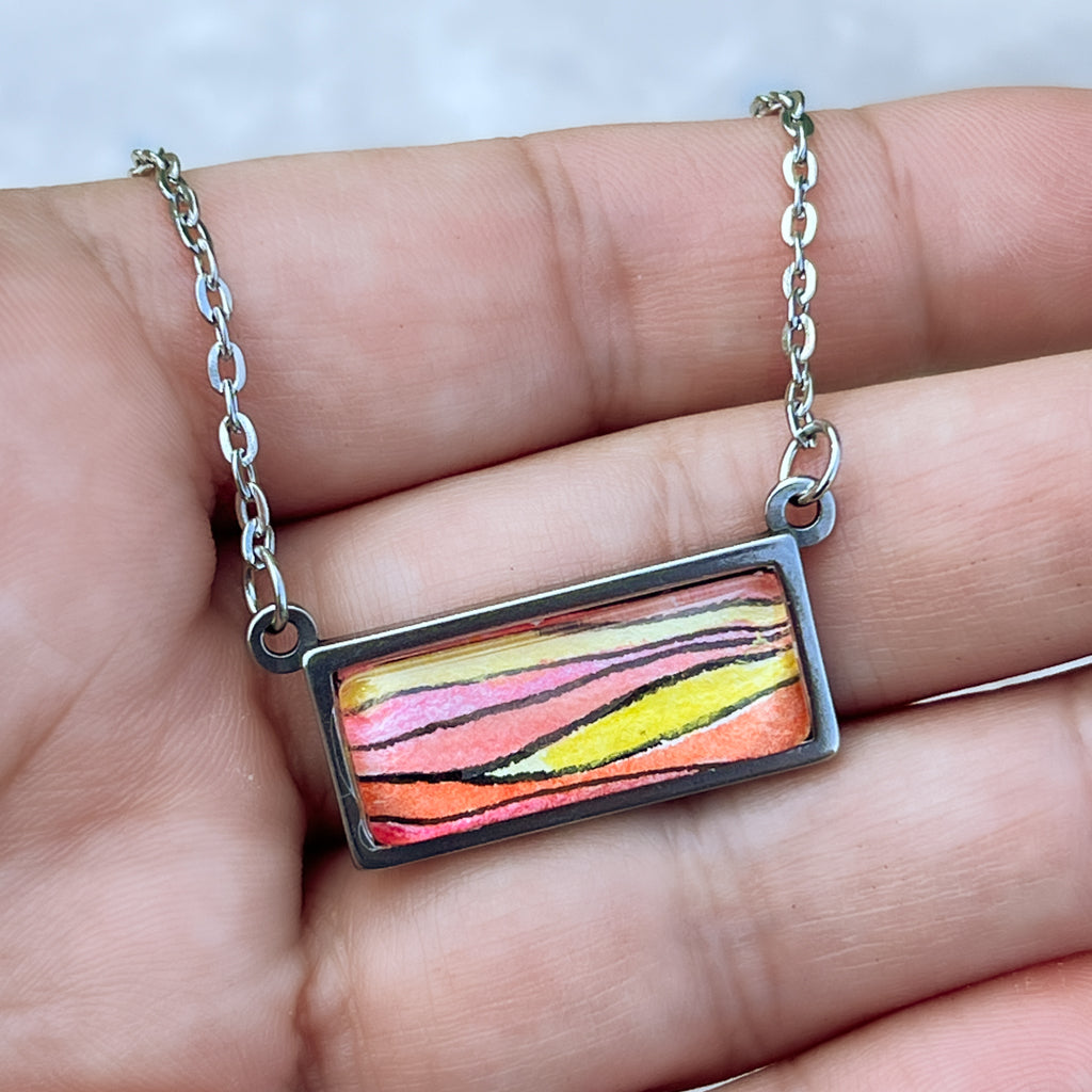 Sunset horizontal necklace shown in hand with detail of orange-hued horizontal waves in watercolor and ink. 
