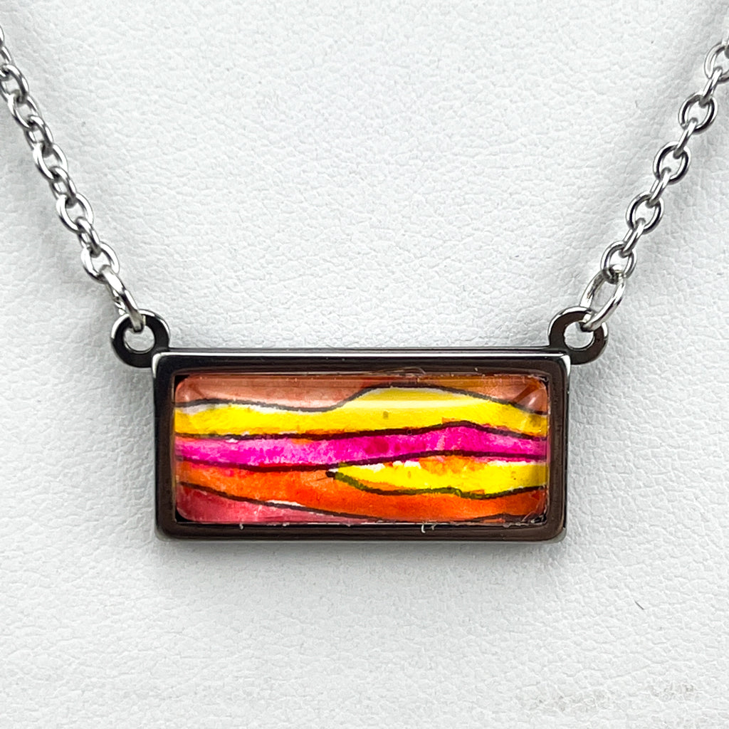 Horizontal stainless steel pendant with a piece of original watercolor art protected behind a glass cabochon.  The art features horizontal waves of pink, orange and yellow.  Closeup on white background. 