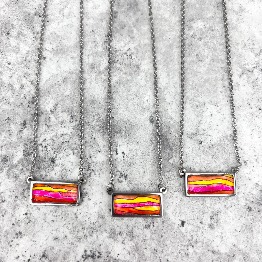 Horizontal stainless steel pendant with a piece of original watercolor art protected behind a glass cabochon.  The art features horizontal waves of pink, orange and yellow.  Closeup of three necklaces showing variation. 