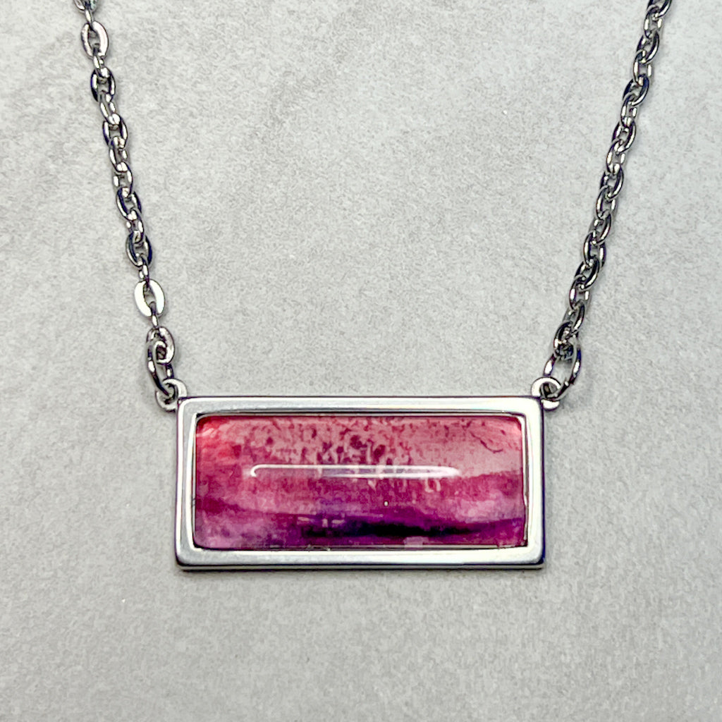 Horizontal rectangular necklace featuring pink and purple watercolor art behind a glass cabochon. 
