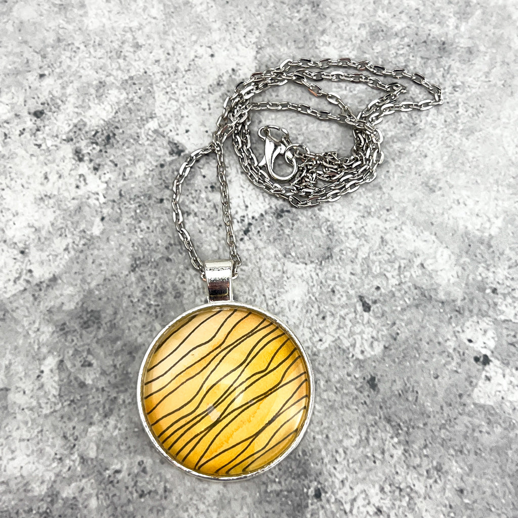 Tiger Stripe round pendant featuring wavy black ink stripes drawn on a yellow watercolor background. 