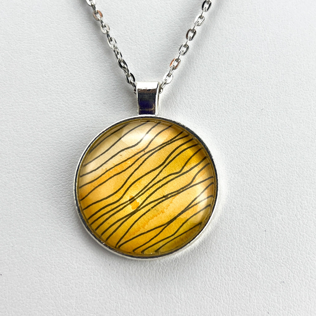 Tiger Stripe round pendant featuring wavy black ink stripes drawn on a yellow watercolor background.  Displayed on a white background. 