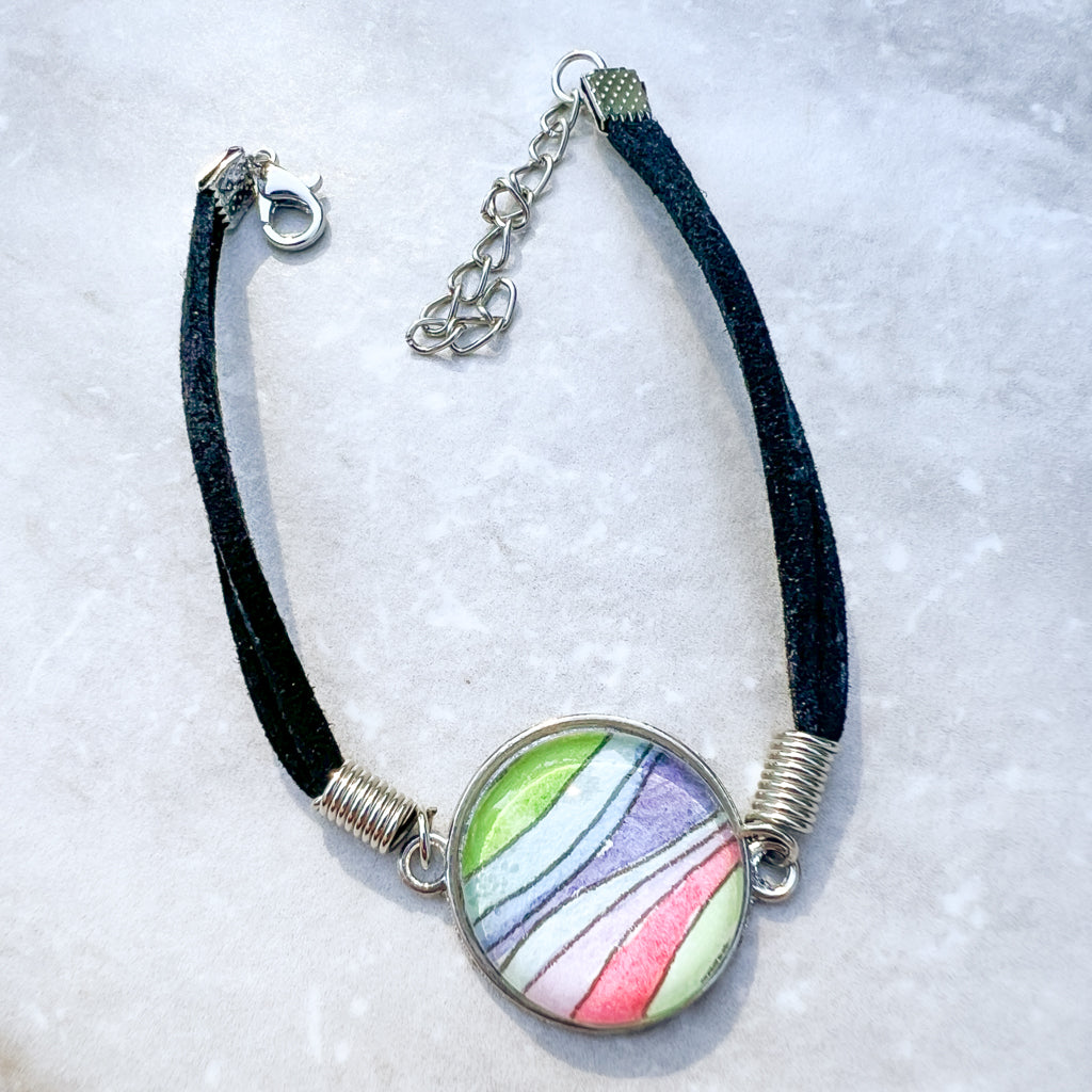 Close up of rainbow-hued watercolor waves set behind a glass cabochon and strung on a black leather bracelet. 