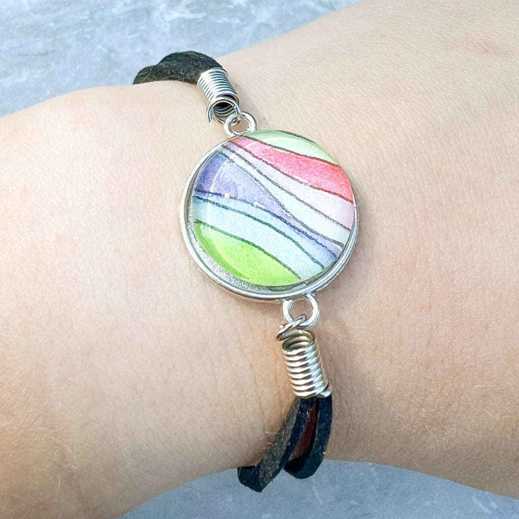 Close up of rainbow-hued watercolor waves set behind a glass cabochon and strung on a black leather bracelet. 