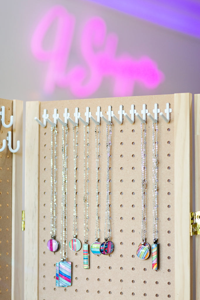Selection of JL Scheppers watercolor necklaces hanging on a peg board underneath a pink neon. 